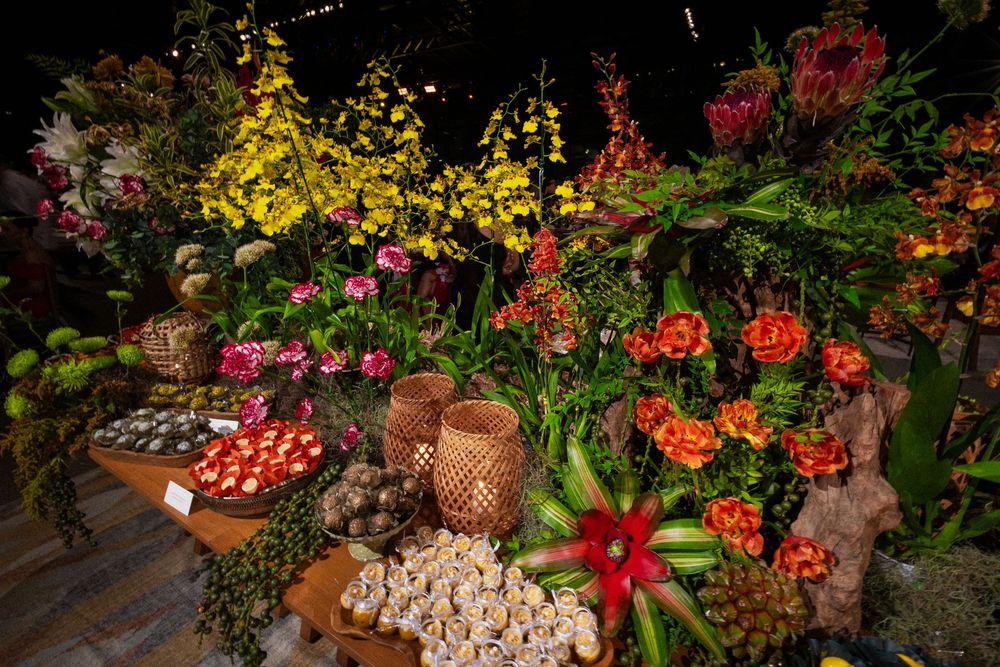 Colorful floral market display of orange blooms, yellow orchids, pink flowers, tropical foliage, and woven baskets with fruits and produce.