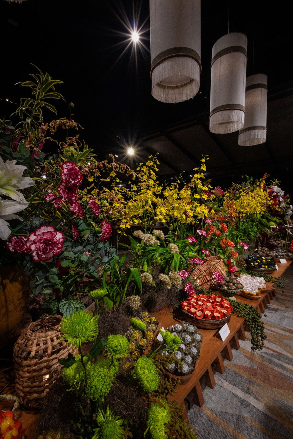 Colorful flower market display with pink peonies, yellow orchids, green chrysanthemums and red fruit baskets under hanging pendant lamps.