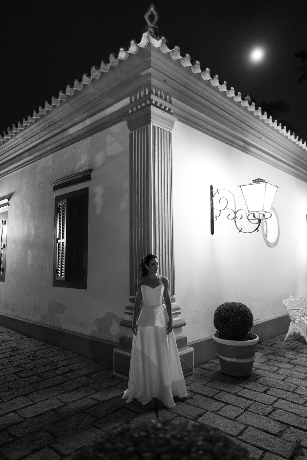 Black and white night photo of a bride in a white gown standing beside a classic building corner under a full moon on a cobblestone patio.