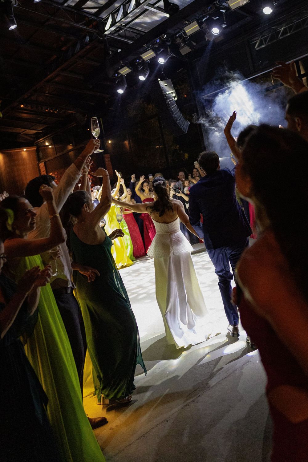 Guests in colorful evening gowns dance with raised arms on a lit dance floor at a lively party under stage lights and haze.