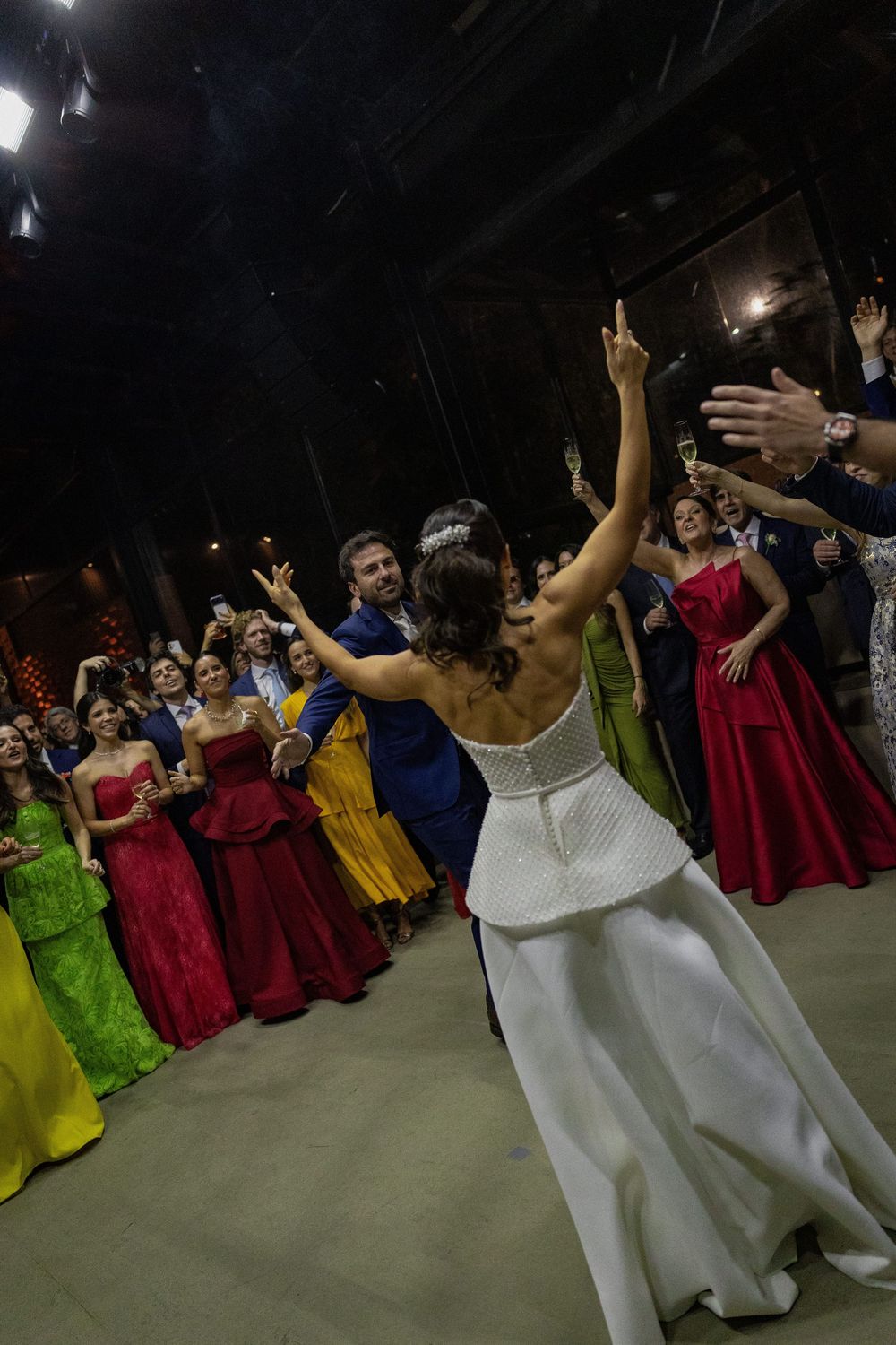 Bride in a white gown dances with arms raised as colorful dressed wedding guests cheer and clap around her in a dimly lit venue.