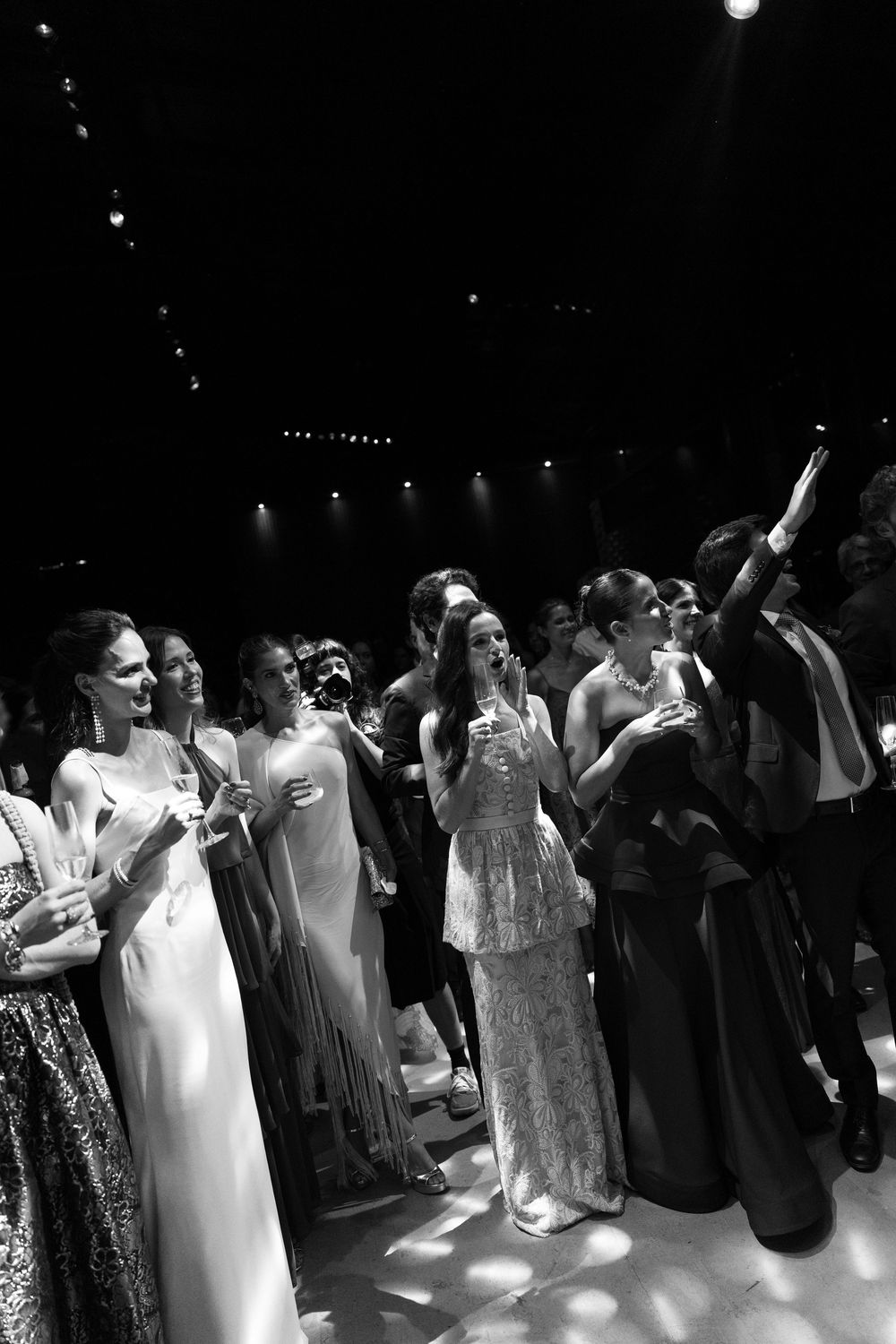 Black and white photo of elegantly dressed guests in gowns and suits raising glasses and gazing upward at a lively evening party.