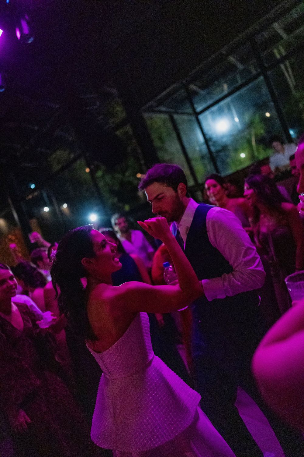 A couple in formal attire dances joyfully at a lively evening reception, surrounded by guests under purple lighting.