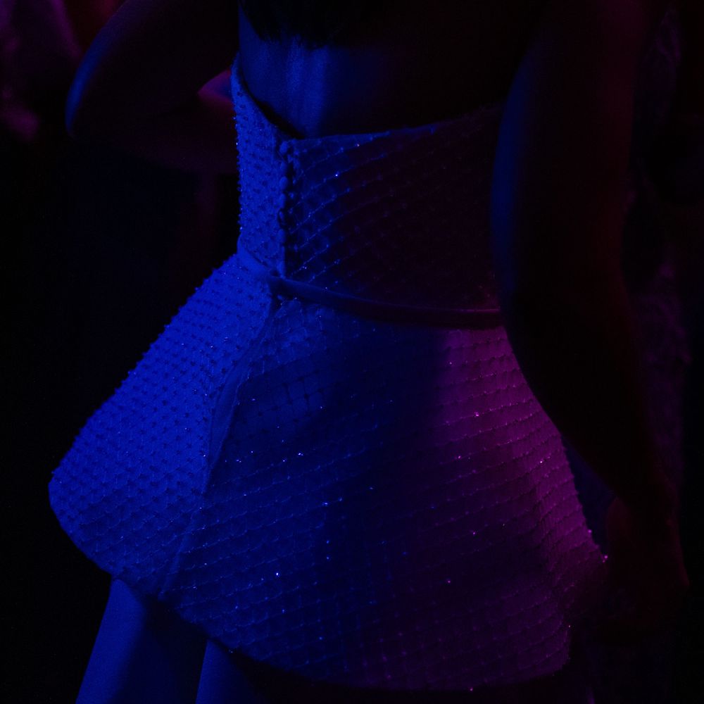 Person in a sparkly white peplum-style dress with a structured flared waist, softly lit by moody blue and purple lighting.