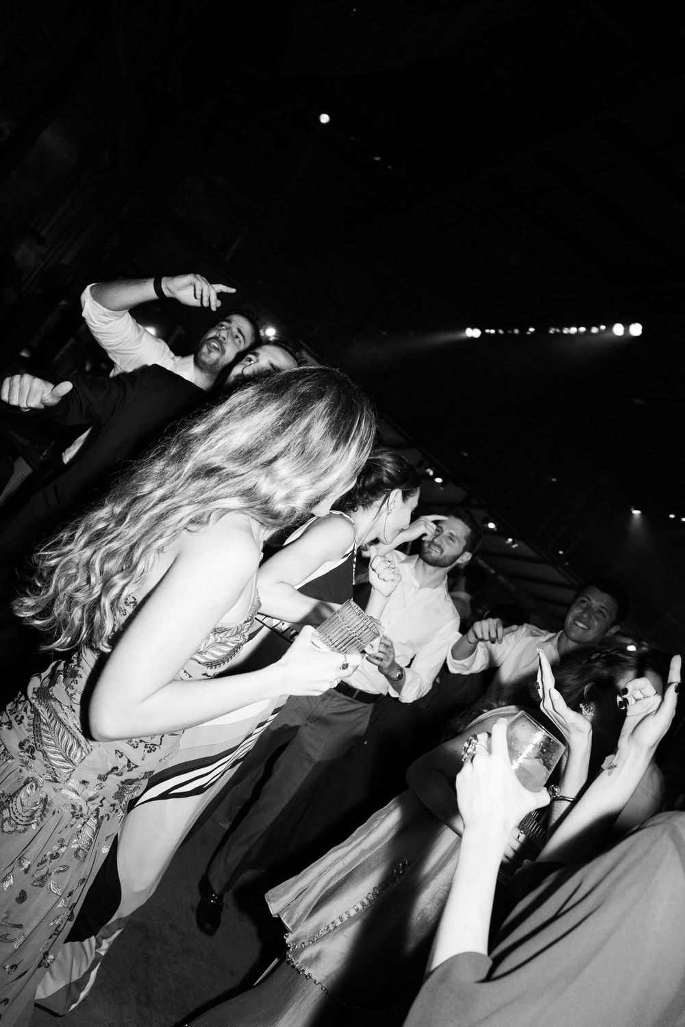 Black and white photo of wedding guests dancing and celebrating together on a crowded dance floor under dramatic stage lighting.