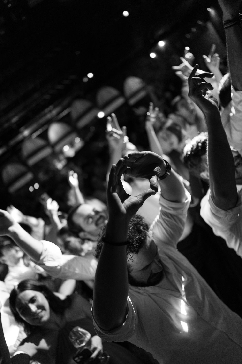 Black and white photo of a cheering crowd at a lively party, hands raised in the air, with one man forming a heart shape above his head.