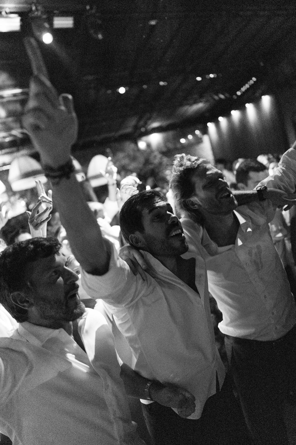 Black and white photo of three men in white shirts dancing and pointing upward at a lively indoor wedding party.