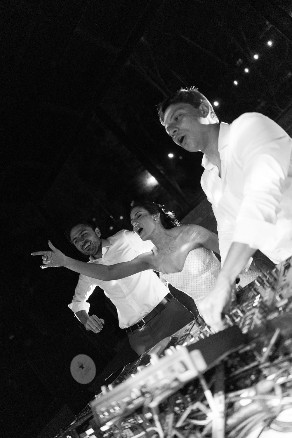 Black and white photo of a DJ in headphones mixing at a turntable while a woman and another man cheer and point on the dance floor.