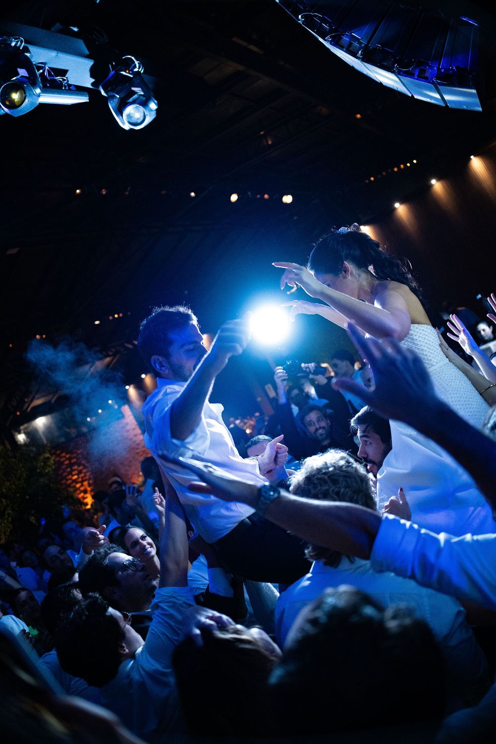 Cheering guests lift two people above the crowd on a dark dance floor lit by bright stage lights and a glowing spotlight at a lively party.