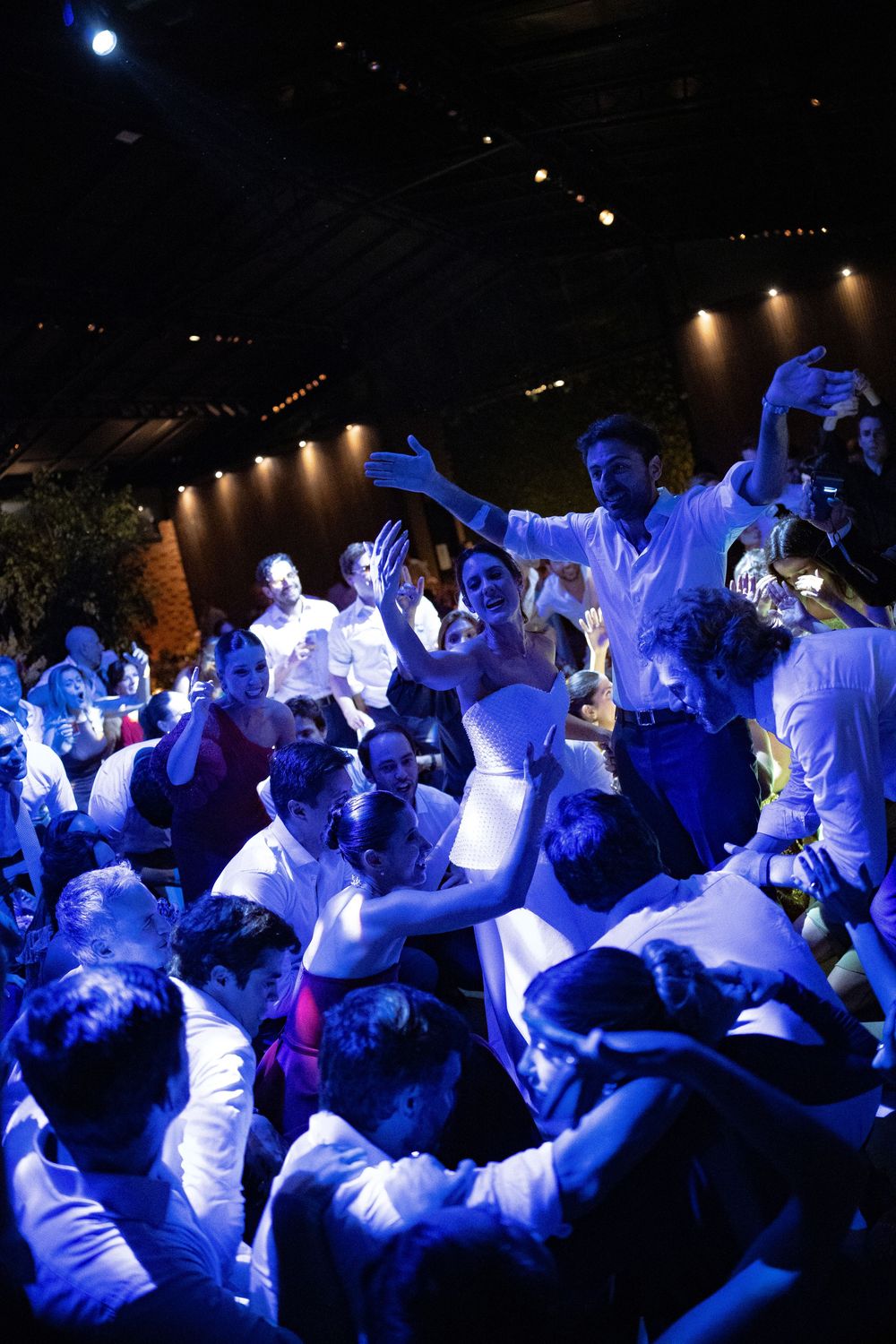Guests in white shirts dance and cheer with raised arms under blue lighting at a lively wedding reception celebration.