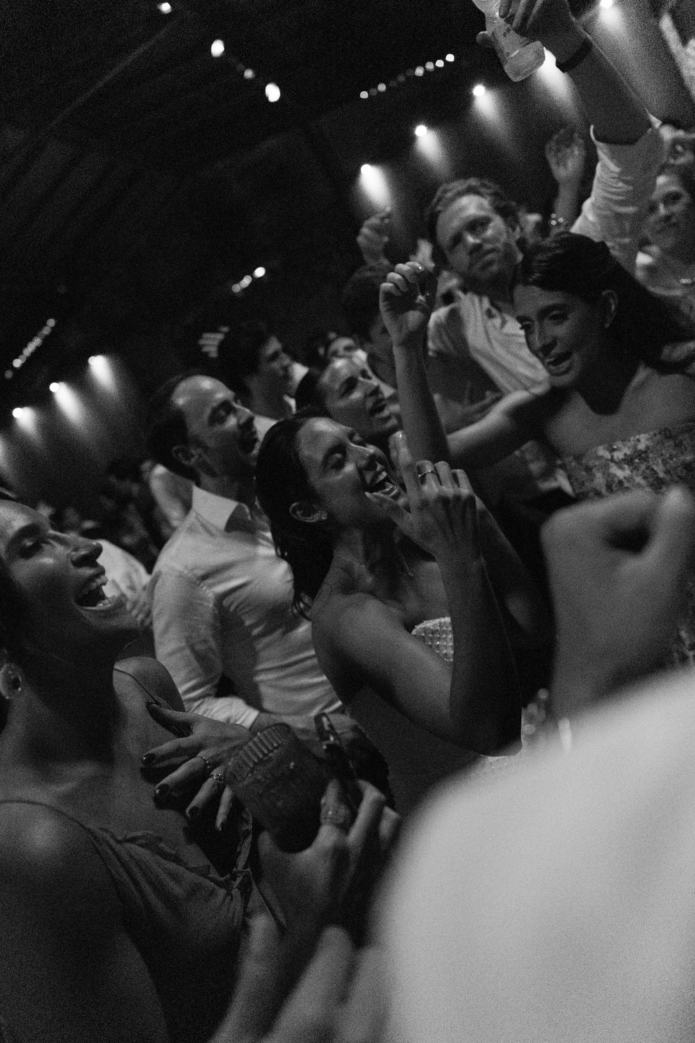 Black and white photo of a lively crowd dancing and singing at a party, with arms raised and warm stage lights glowing overhead.