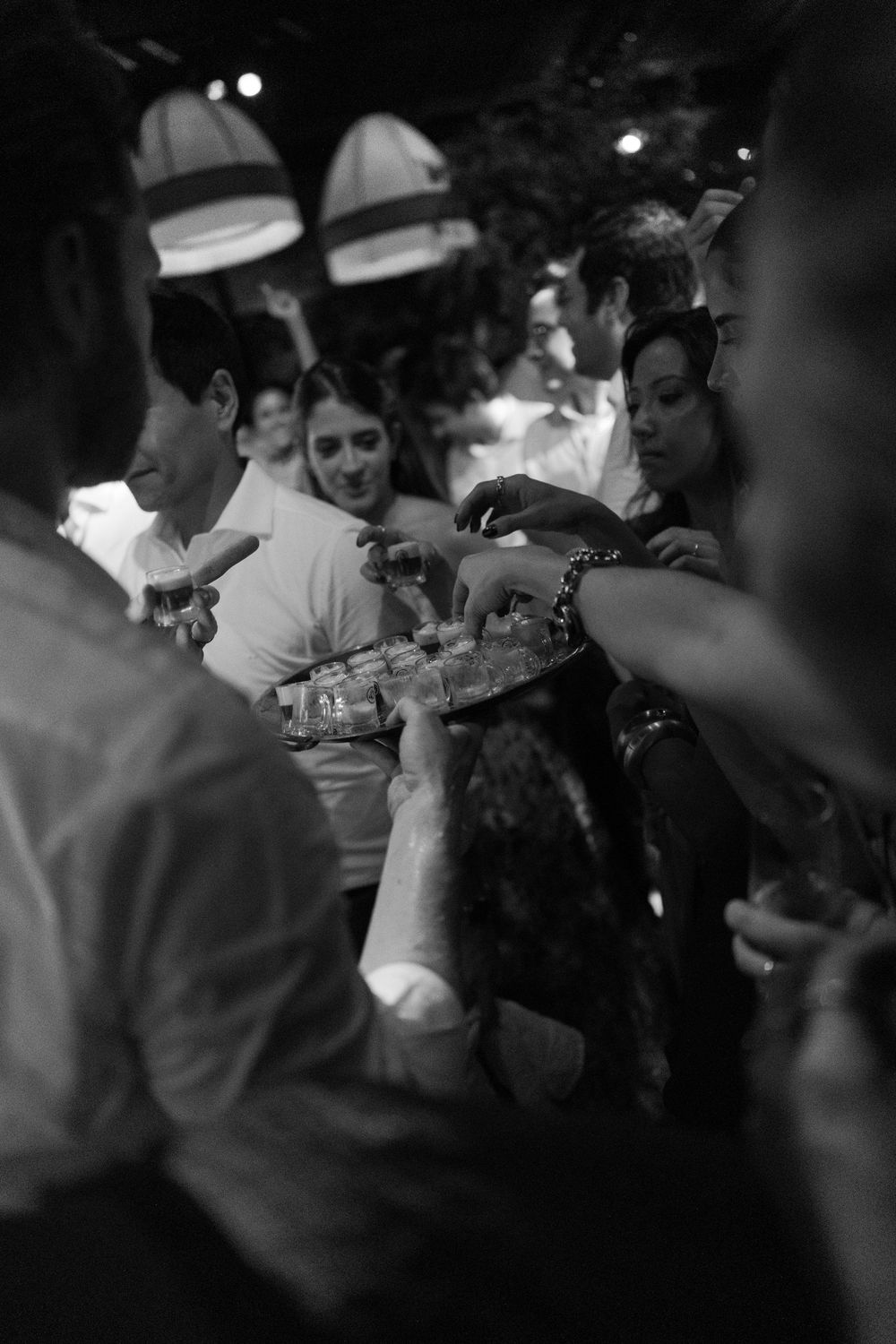 Black and white photo of guests at a lively gathering reaching for shot glasses on a tray under warm pendant lights.