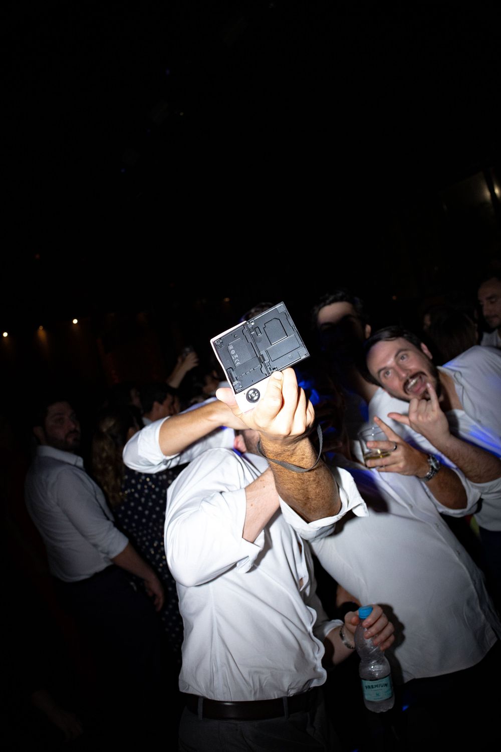Group of men in white shirts celebrating at a dark party while one holds a camera up to take a selfie and another gestures.