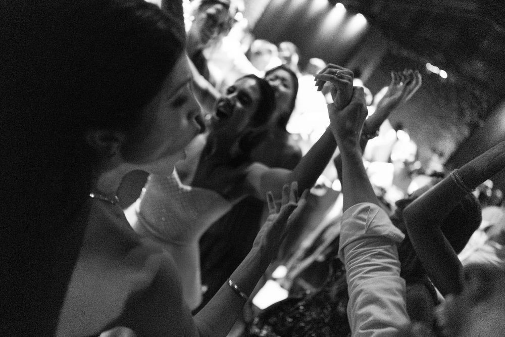 Black and white photo of a lively crowd dancing at a celebration, with guests raising their hands in the air under warm stage lights.