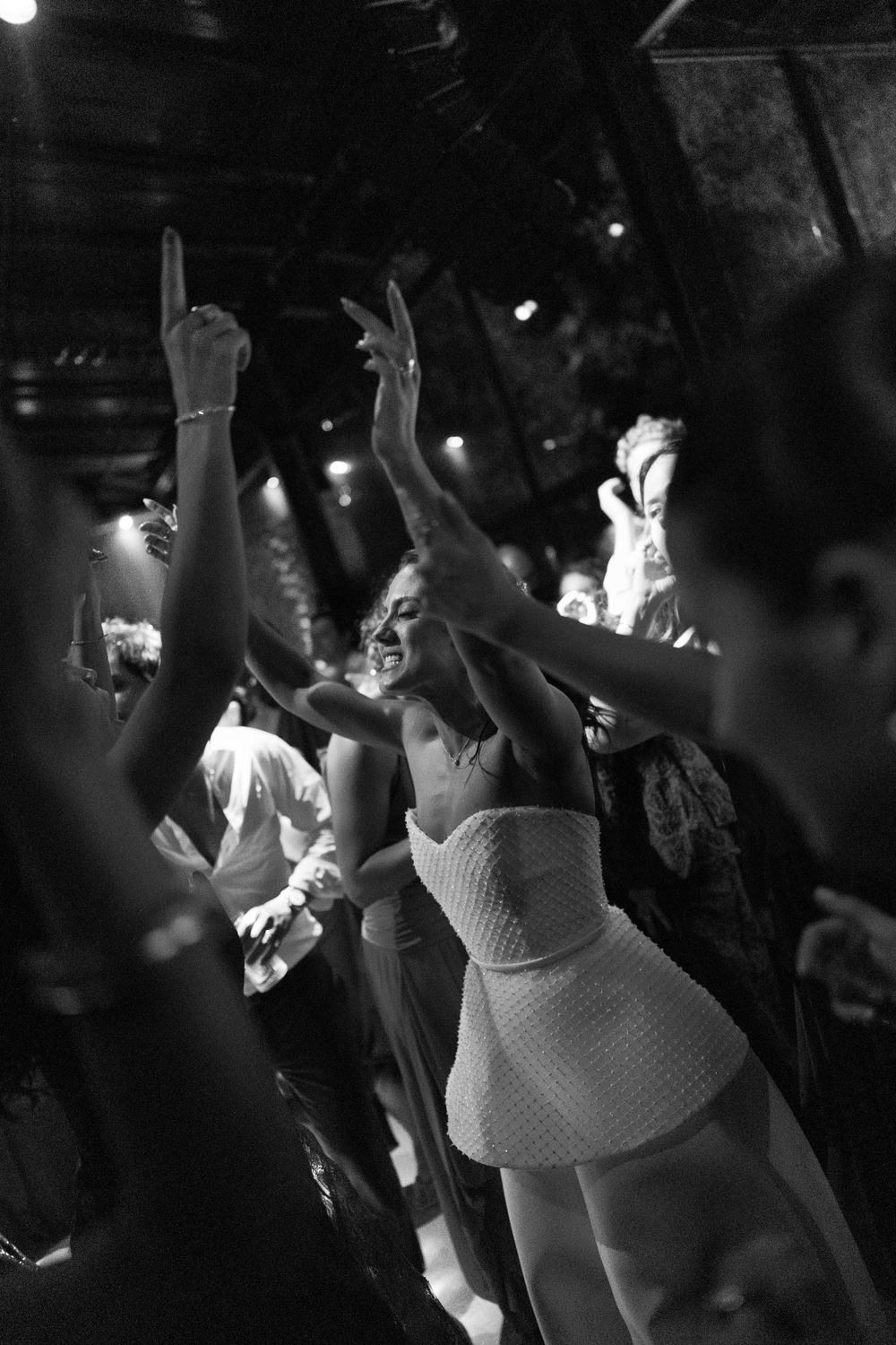 Black and white photo of a smiling woman in a white dress dancing with arms raised among cheering guests at a lively evening celebration.