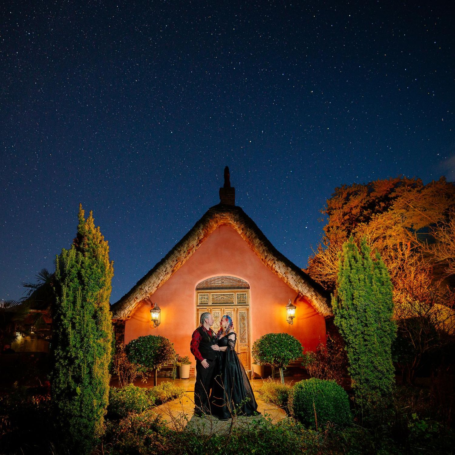 Bride and groom embracing outside the pink thatched cottage at Le Petit Chateau, Otterburn, under a starry night sky on their Halloween wedd