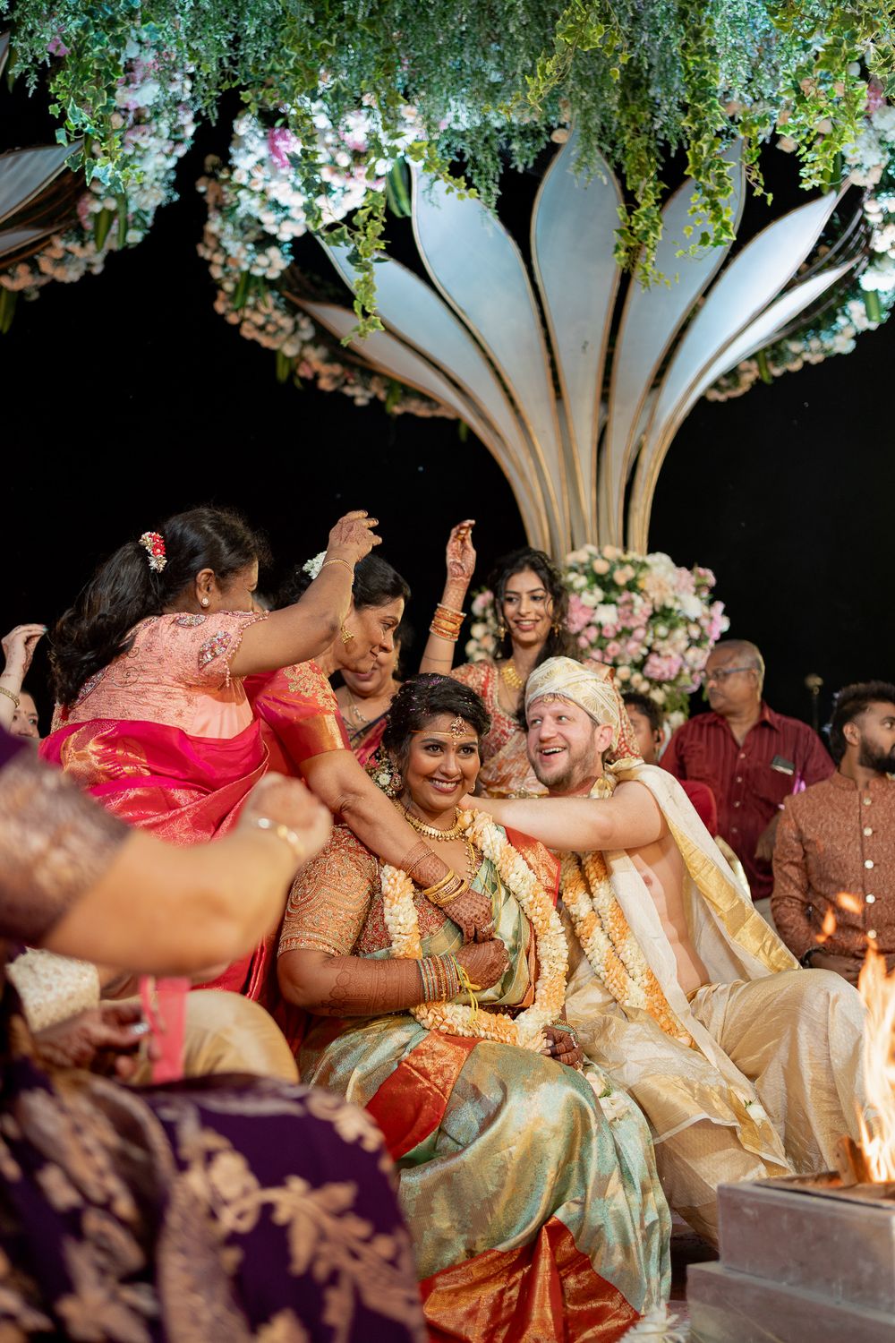 Joyful Indian wedding ceremony with bride in gold saree and groom in cream sherwani surrounded by celebrating guests under floral decor.