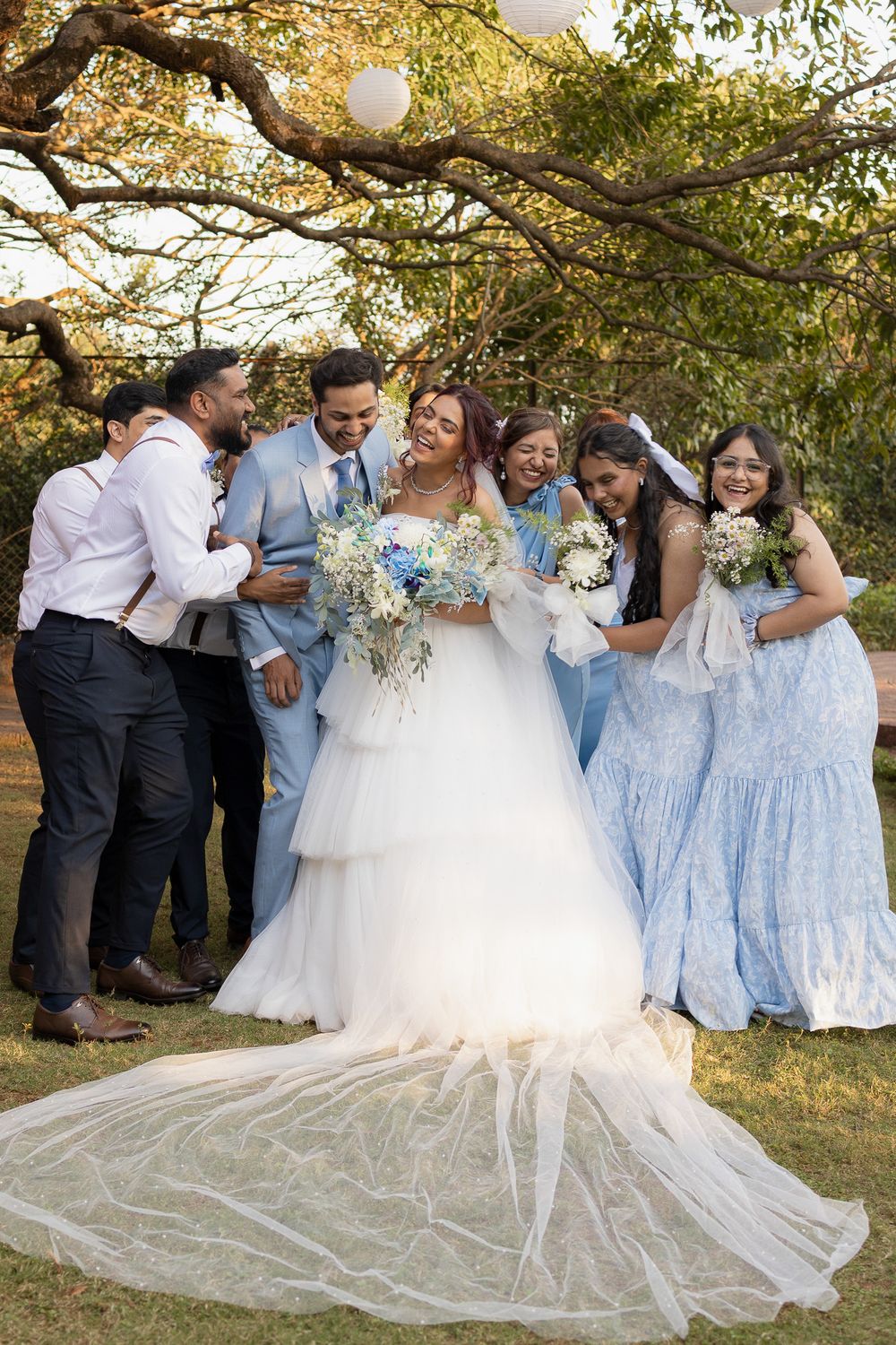 Bride in white gown with bridal party in blue dresses and suits celebrating outdoors under trees.