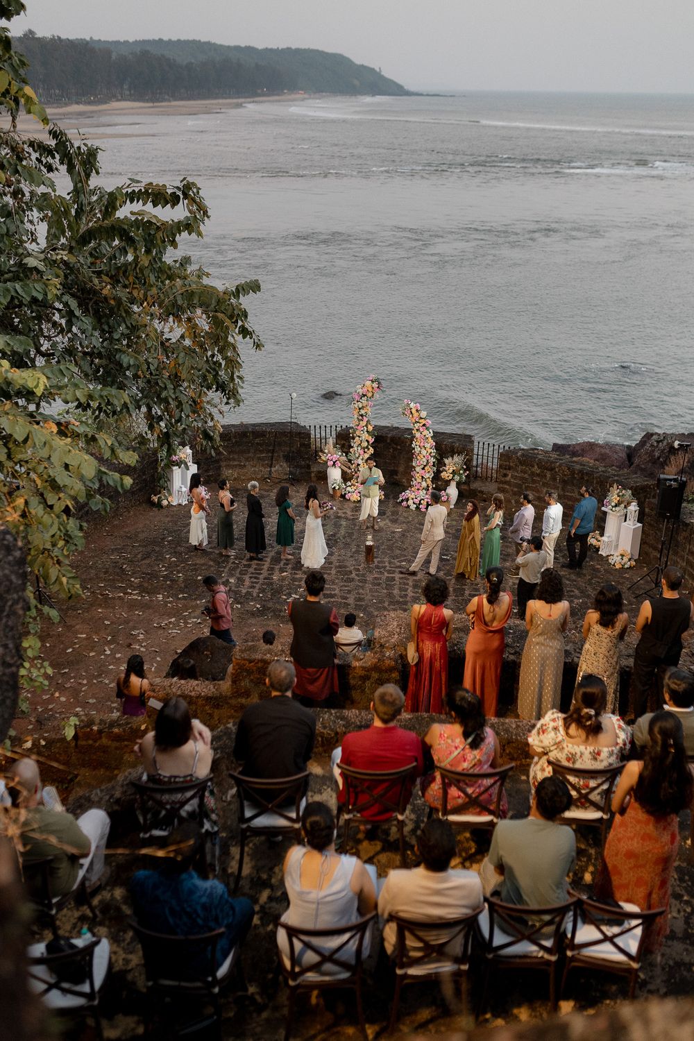 Aerial view of an intimate outdoor wedding ceremony on a coastal cliff overlooking the ocean, with guests in colorful attire.