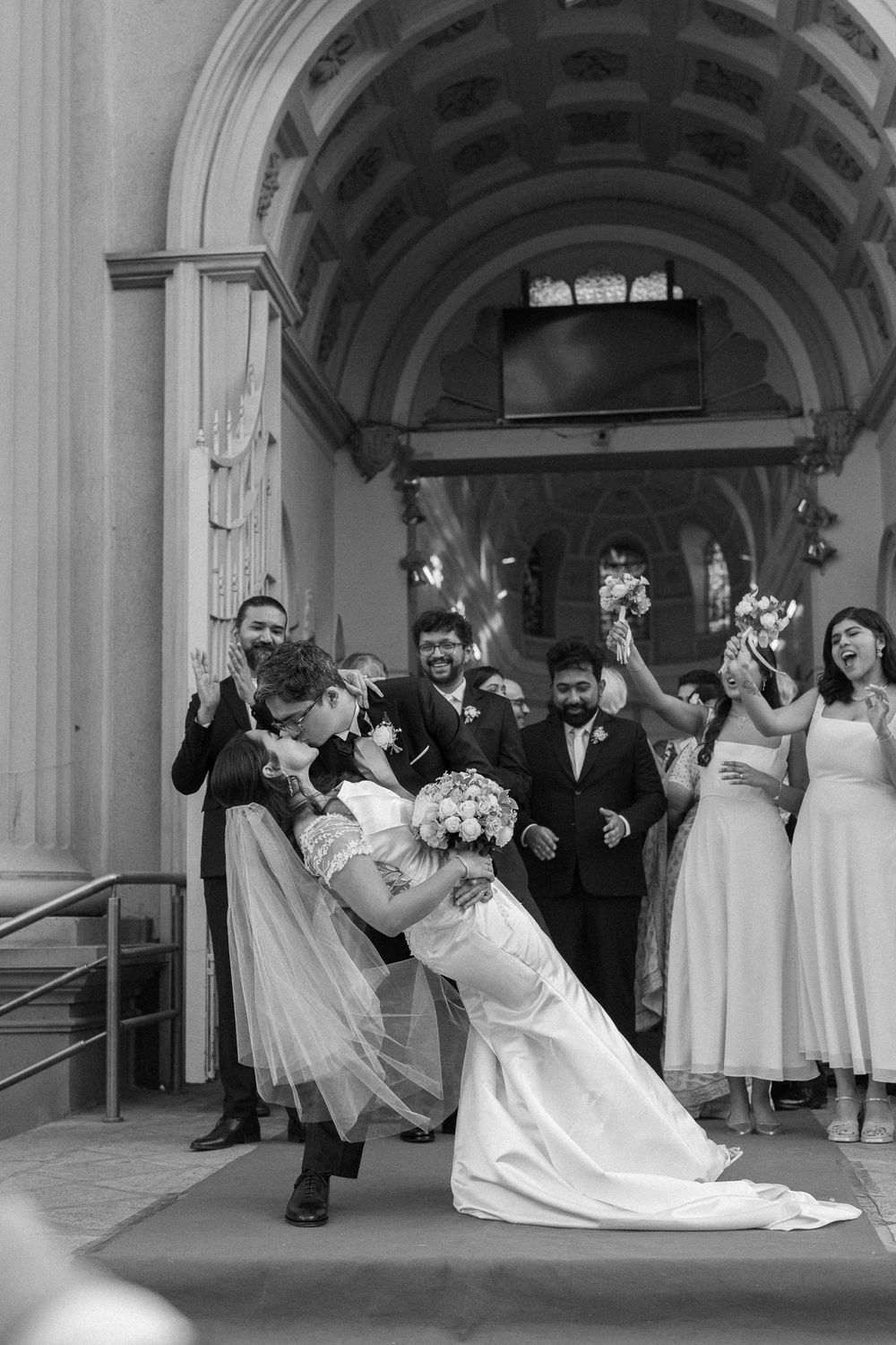 Black and white photo of newlyweds sharing a romantic dip kiss outside a church surrounded by cheering wedding party.
