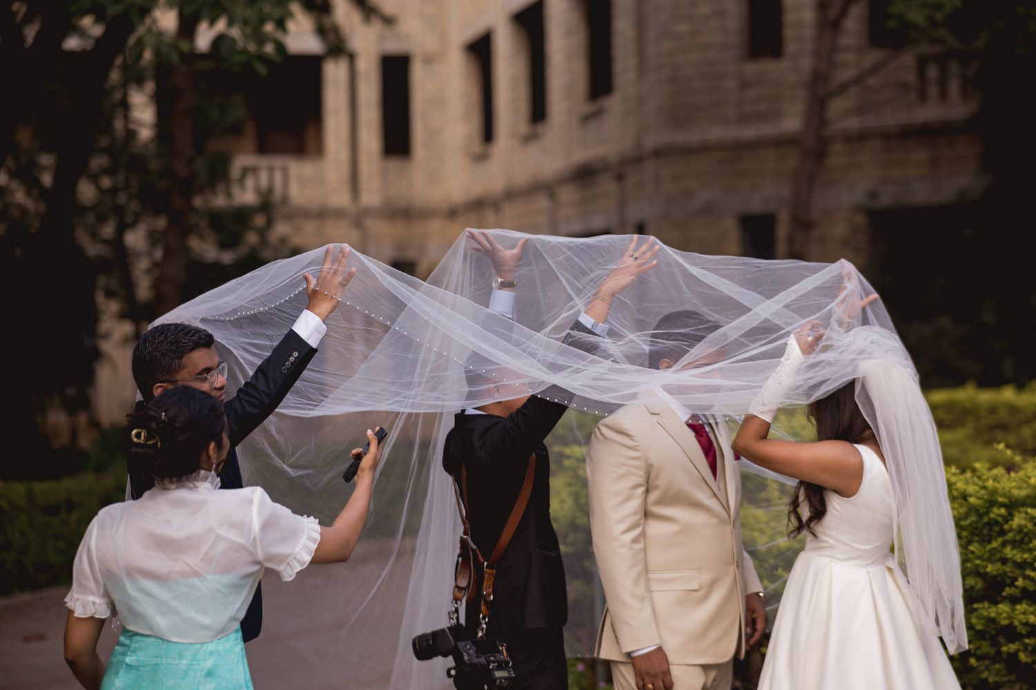 Wedding party holds bride's veil overhead outdoors, with photographer capturing the romantic bridal moment.