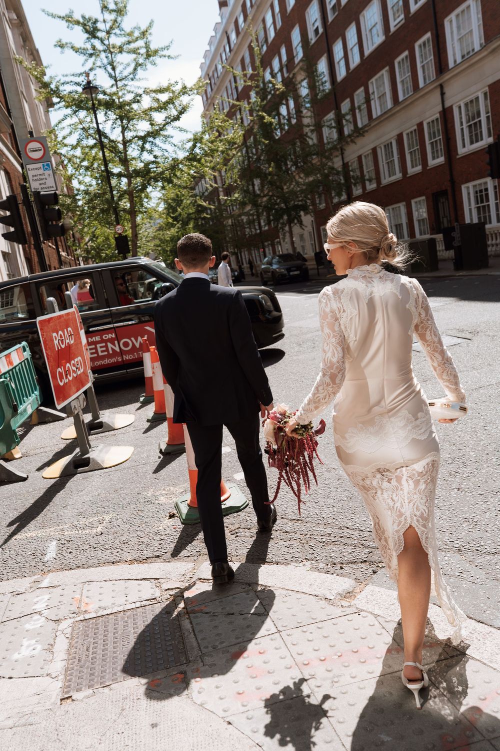 Bride in lace dress and groom in black suit walk hand-in-hand past road closed signs on a sunny London street.