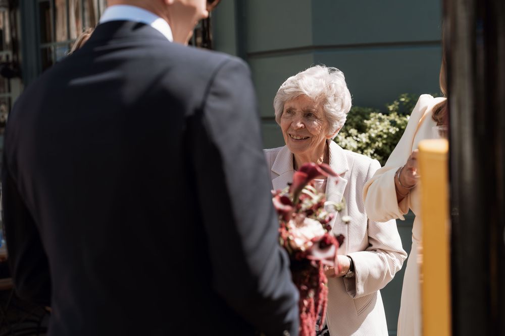 Elderly woman in pink jacket smiling warmly at a man in a dark suit outdoors at a formal event.
