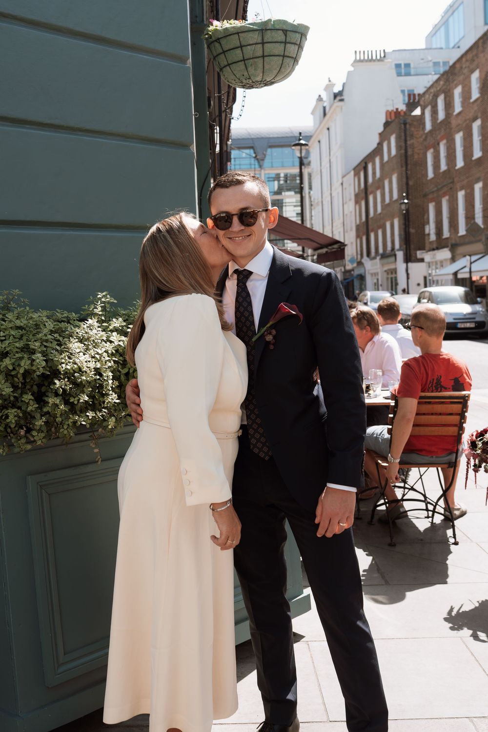A woman in a white dress kisses a man in a dark suit on the cheek outside a street cafe on a sunny day.