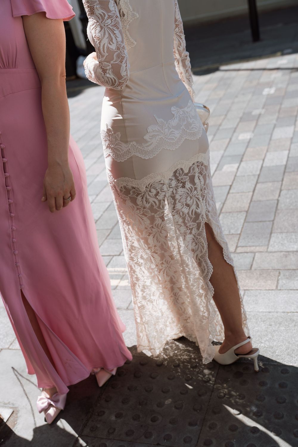 Bride in lace wedding dress and bridesmaid in pink gown walking on cobblestone pavement.