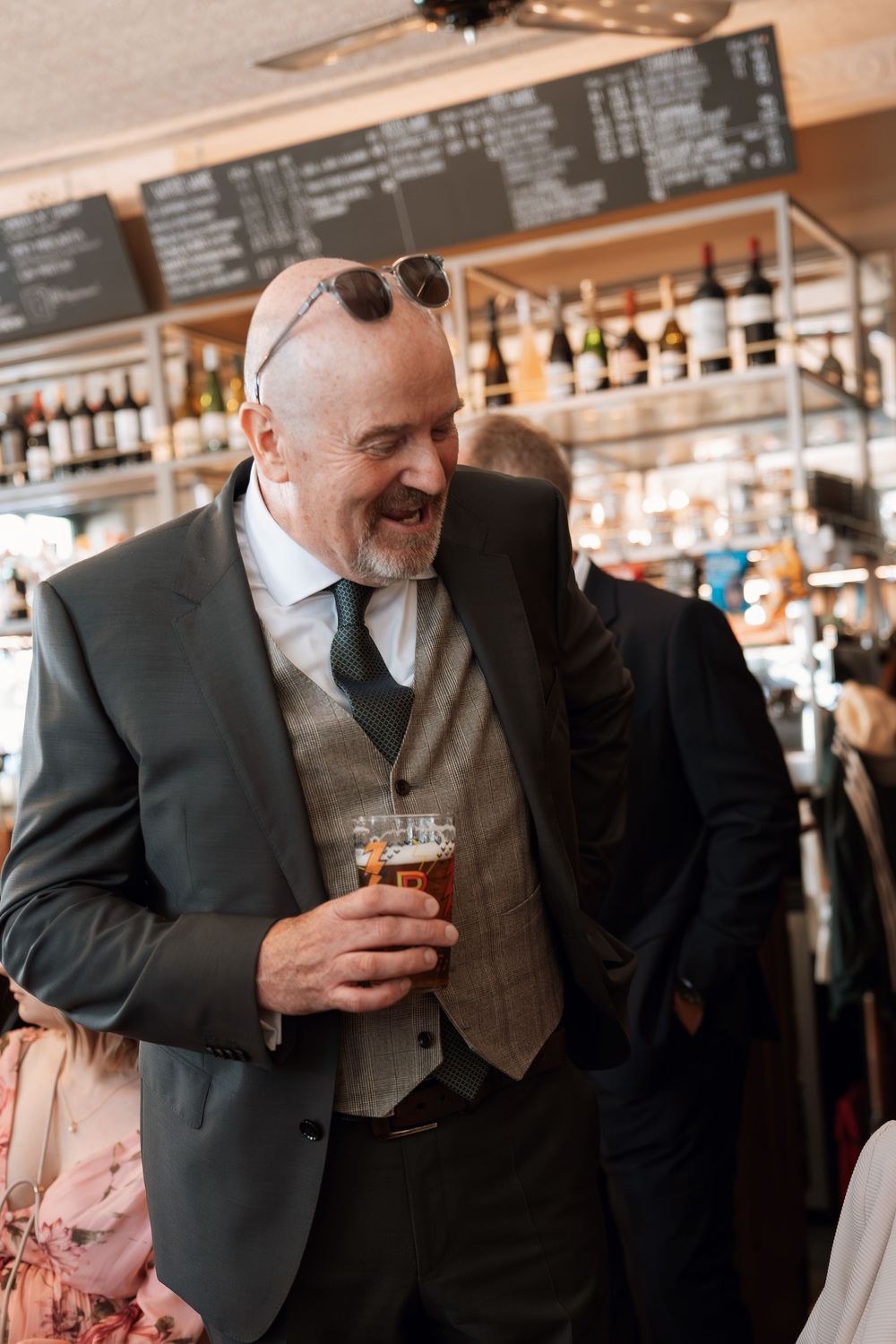 Smiling man in suit and waistcoat holding a pint of beer at a bar, wearing sunglasses on his head.