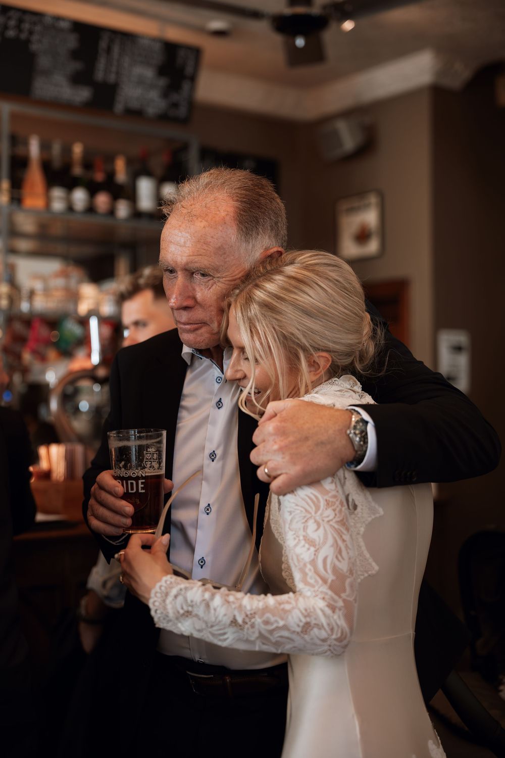 Bride in lace wedding dress hugging older man holding beer pint in a warmly lit bar setting.