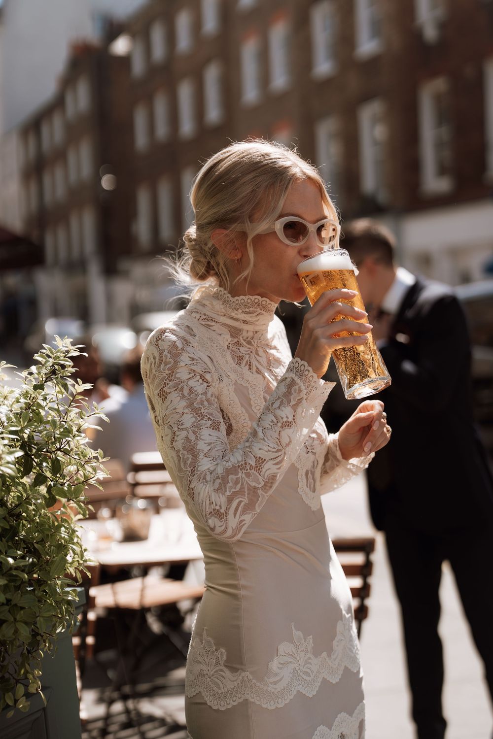 Bride in lace wedding dress drinking a pint of beer outdoors on a sunny city street.
