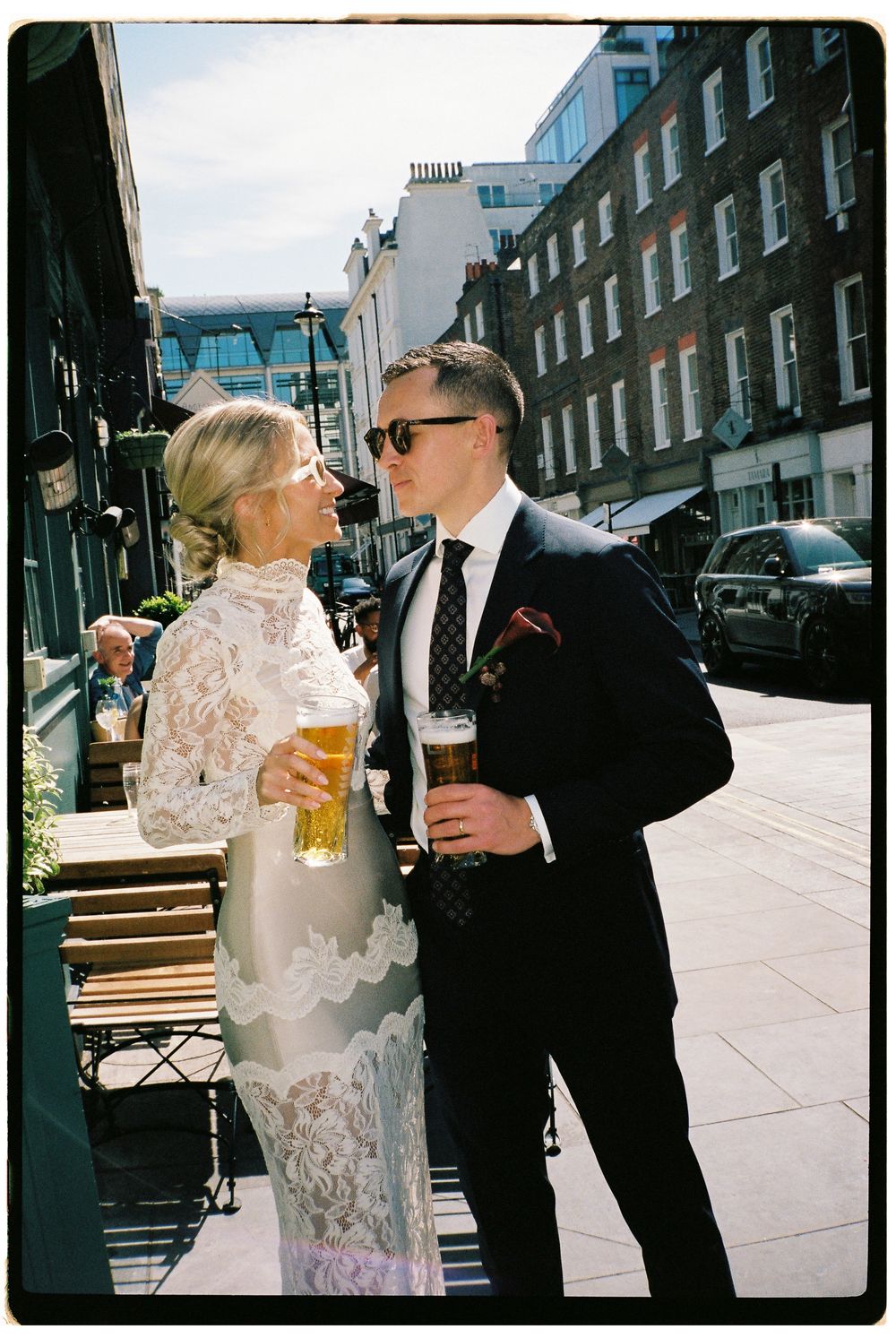Bride in lace gown and groom in dark suit holding beers on a sunny London street.