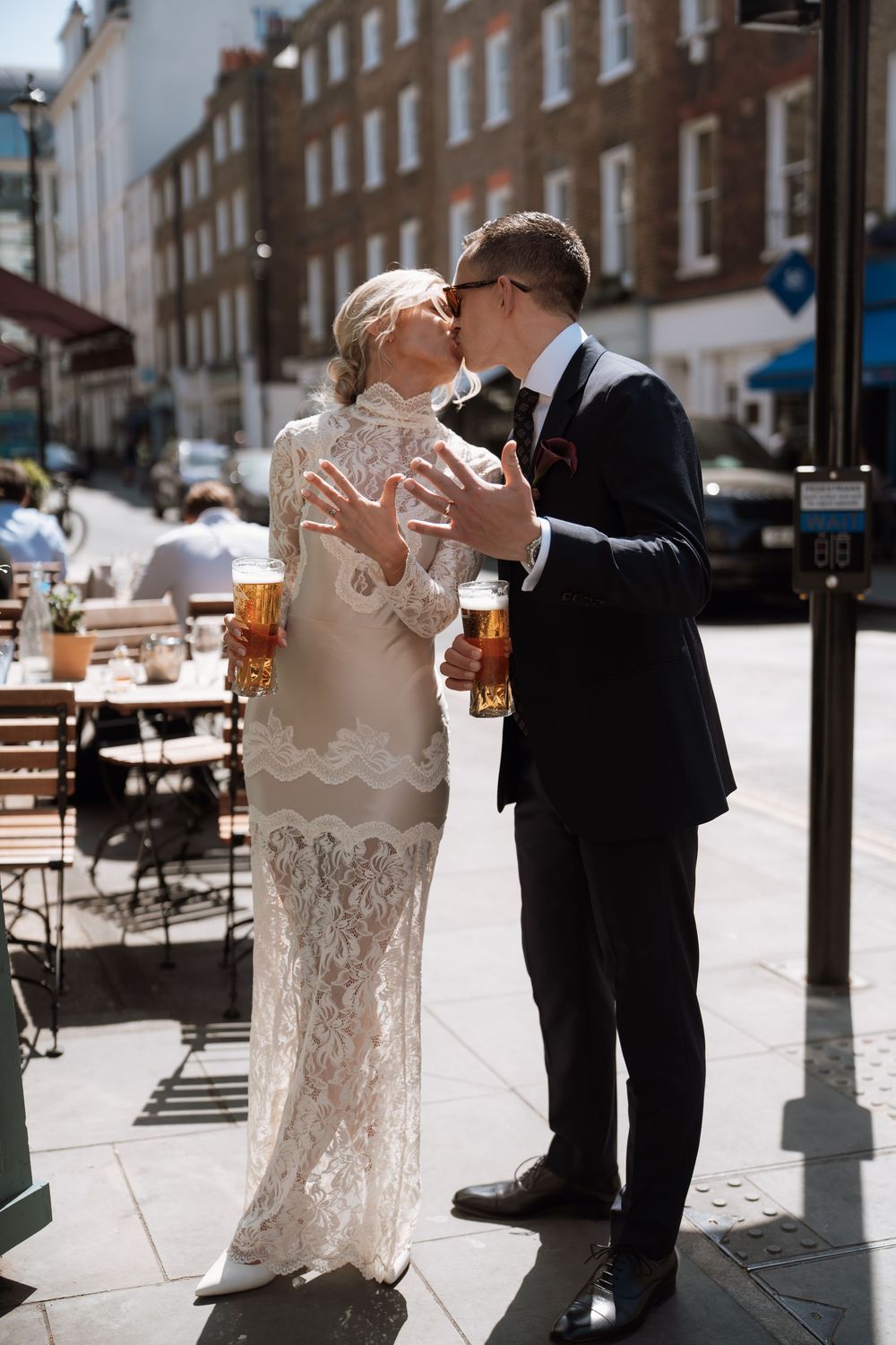 Bride and groom kissing on a sunny London street, holding pints of beer, wearing lace wedding dress and black suit.