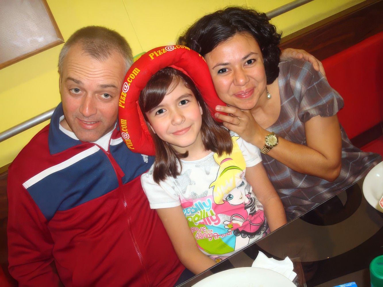 A family of three smiling together, with a young girl wearing a red Flamenco hat between two adults.
