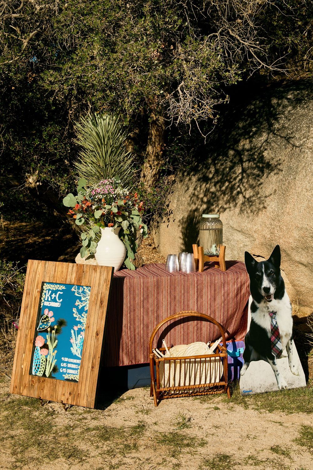 Outdoor wedding setup with a border collie, picnic basket, floral arrangement, and decorative signboard in a natural setting.