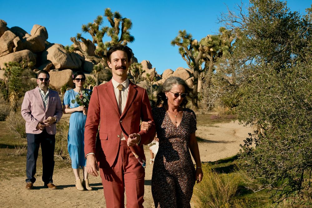 Couple walks hand-in-hand at Joshua Tree desert wedding, guests in background amid boulders and cacti.
