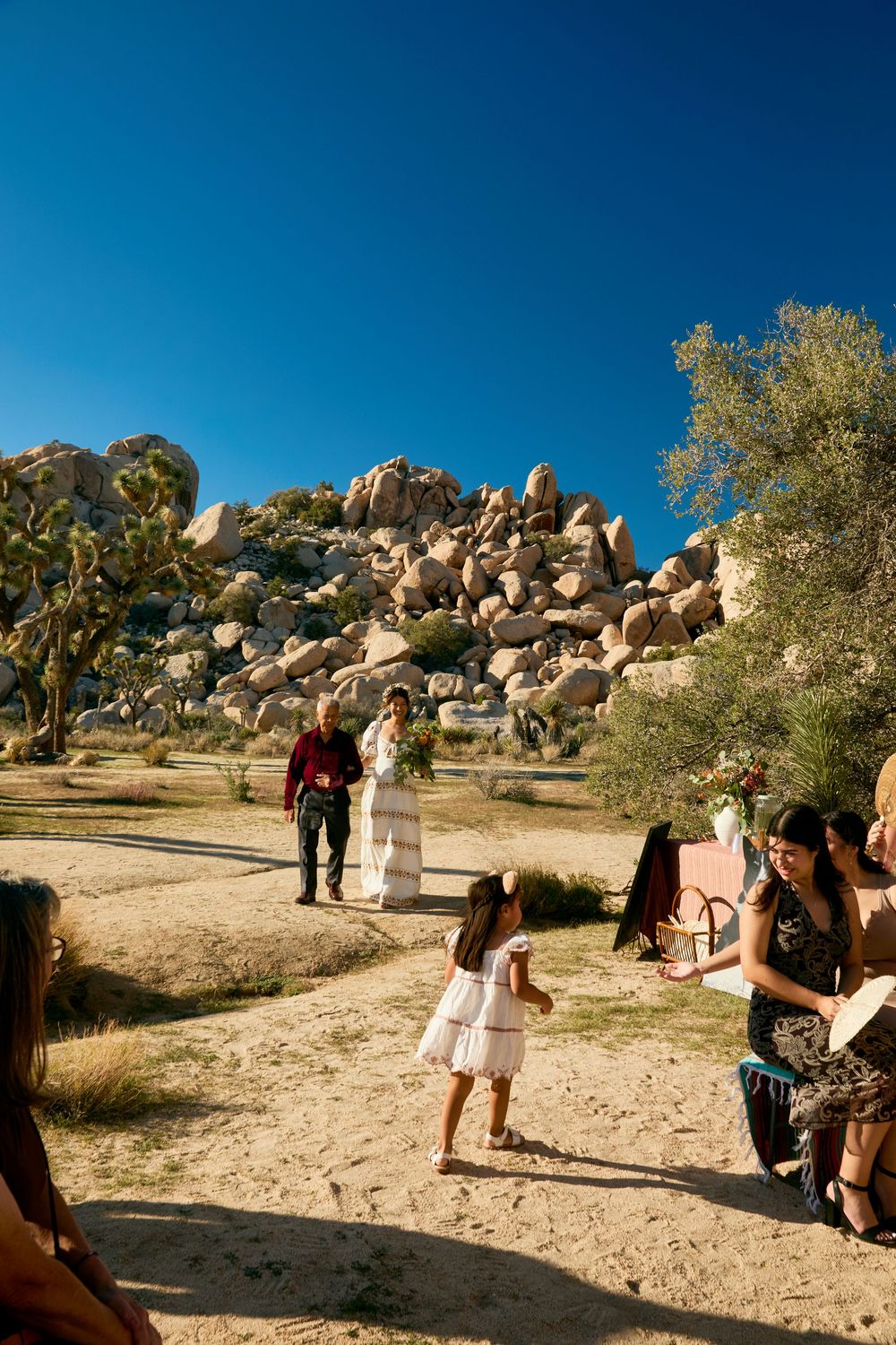 Outdoor desert wedding ceremony at Joshua Tree with guests gathered among boulders and Joshua trees under a clear blue sky.