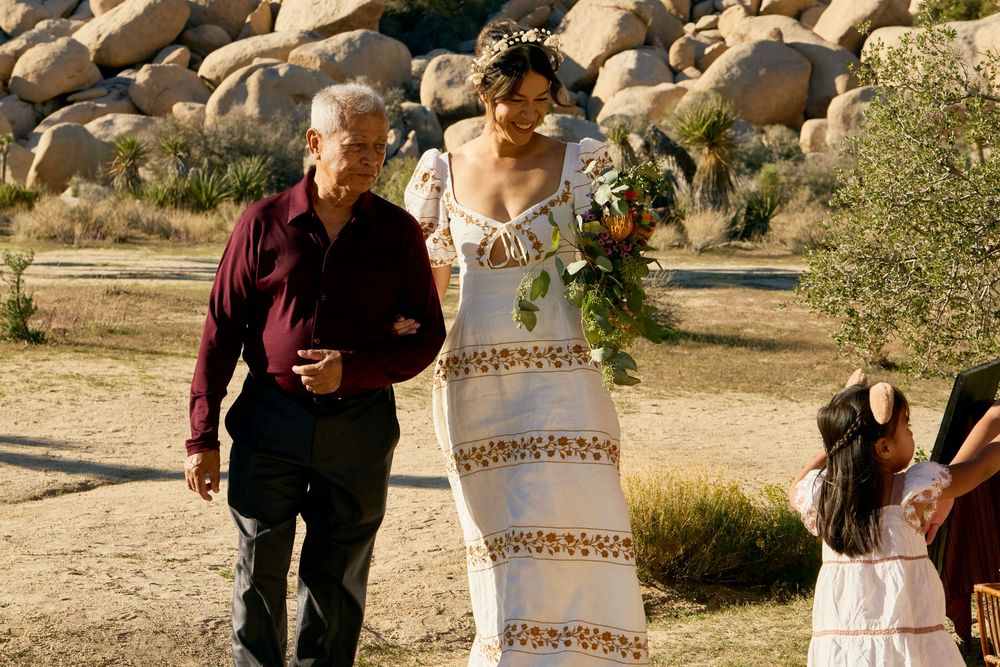 Bride in white floral gown walks with older man through desert landscape with boulders at outdoor wedding ceremony.