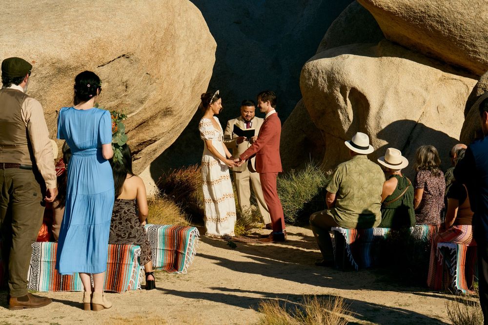 Outdoor desert wedding ceremony among large boulders with guests seated on colorful blankets.