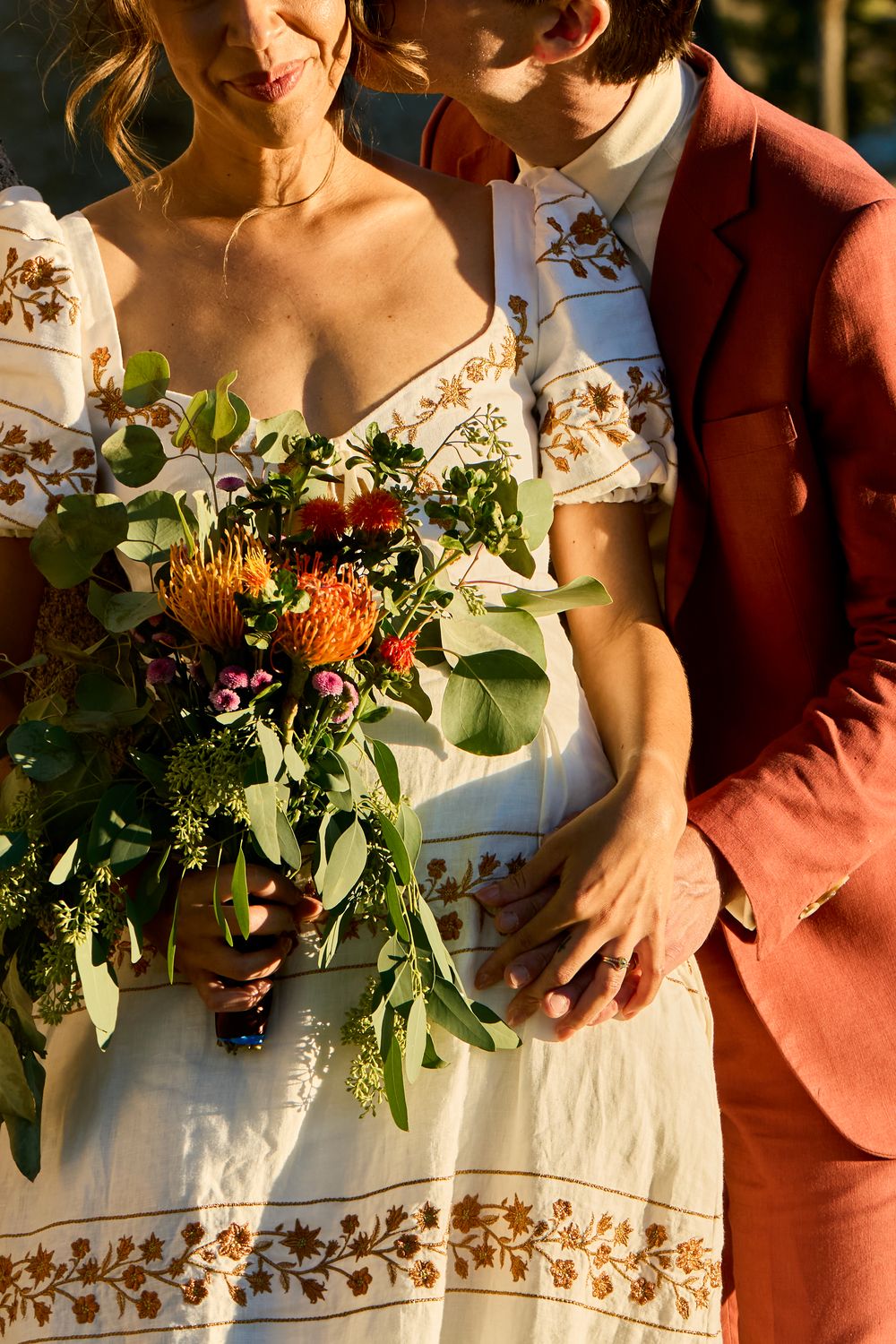 Bride in floral white dress holds vibrant bouquet with greenery, standing beside groom in rust suit at golden hour.