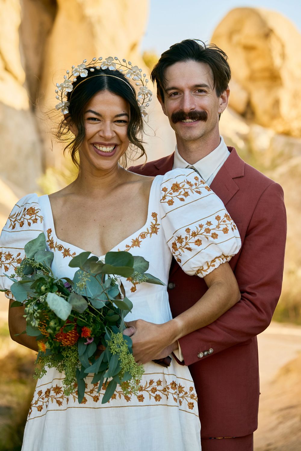 Smiling bride in floral dress with bouquet and groom in rust suit pose together among desert rocks.