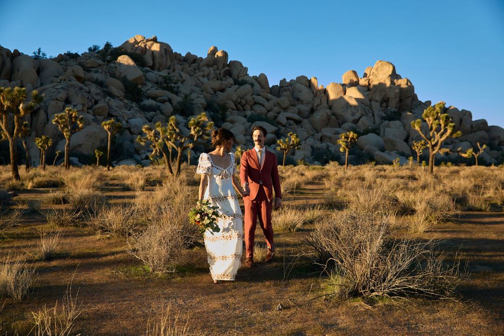 Bride and groom walking hand in hand through Joshua Tree desert landscape with dramatic boulder formations at sunset.