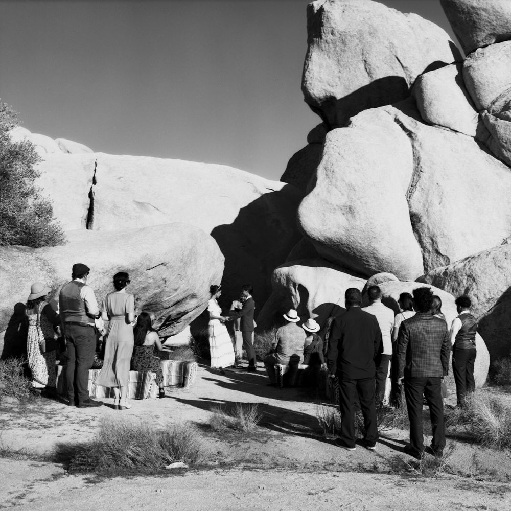 Vintage black and white photo of a crowd gathering near large desert rock formations in arid landscape.