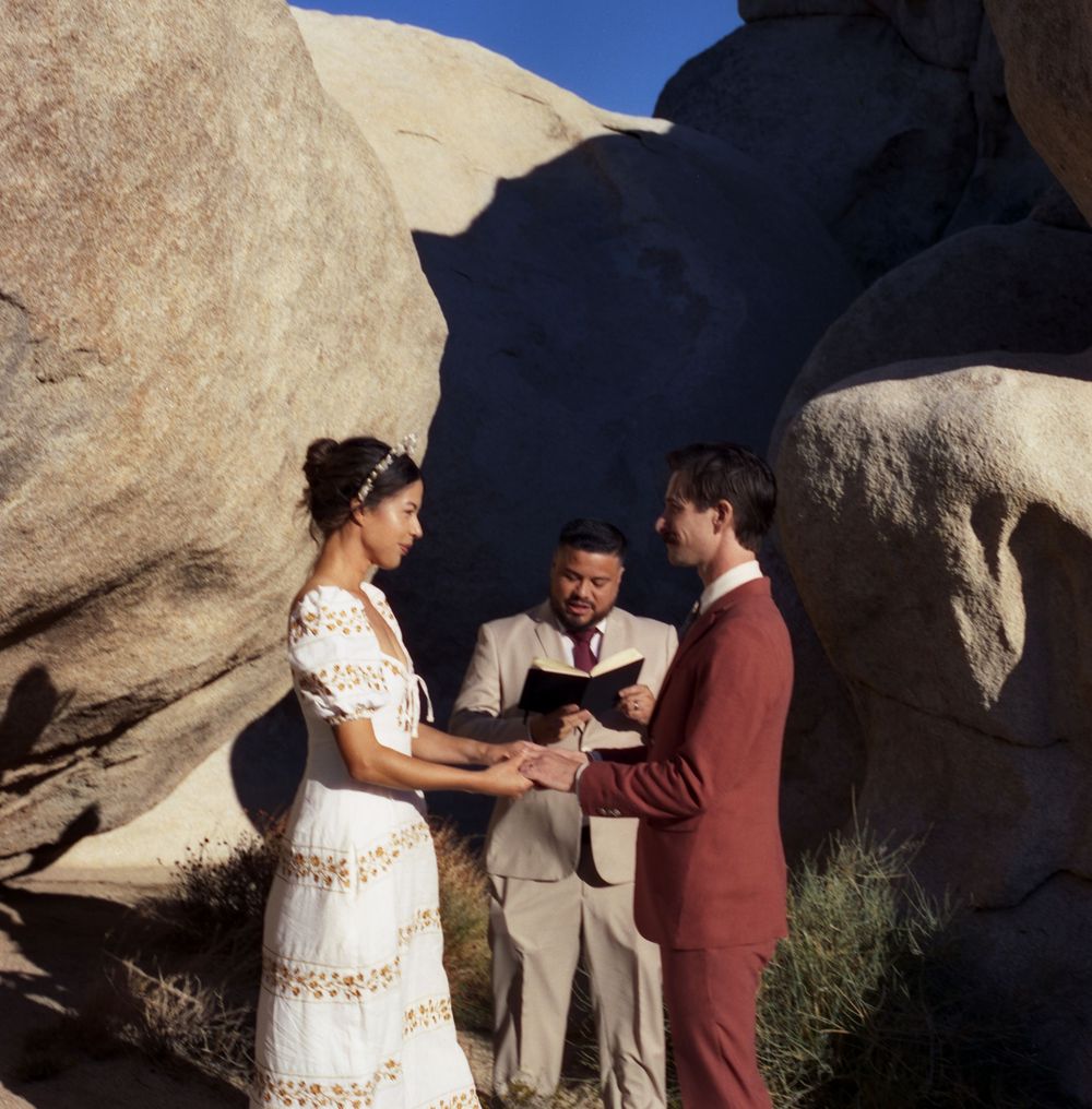 Couple exchanging vows outdoors among large boulders, officiant reading from book, bride in white dress, groom in burgundy suit.