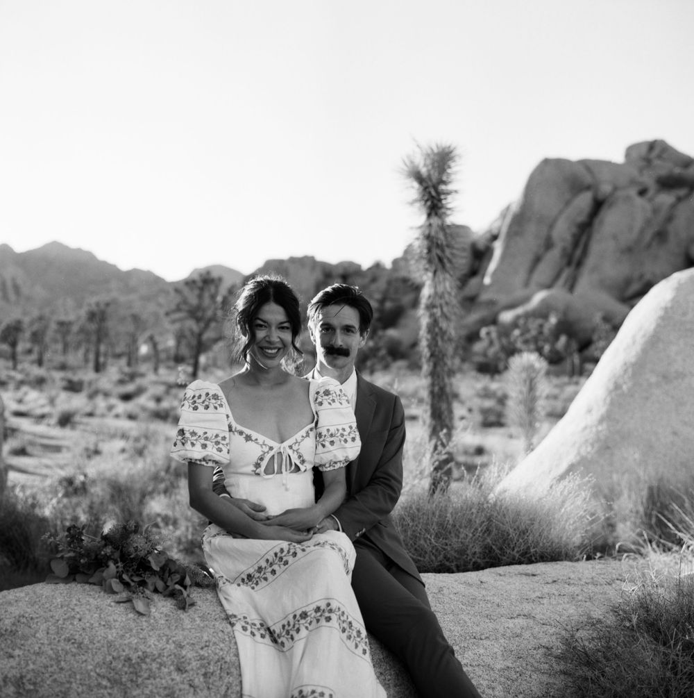 Black and white photo of bride and groom sitting on rocks in Joshua Tree desert landscape.