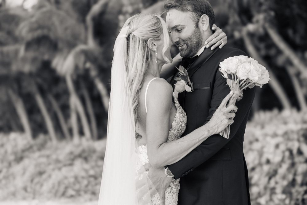 Black and white wedding photo of bride and groom embracing outdoors, bride holding bouquet, palm trees in background.