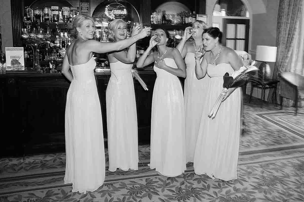 Black and white photo of four bridesmaids in long white gowns laughing and celebrating together at a bar.