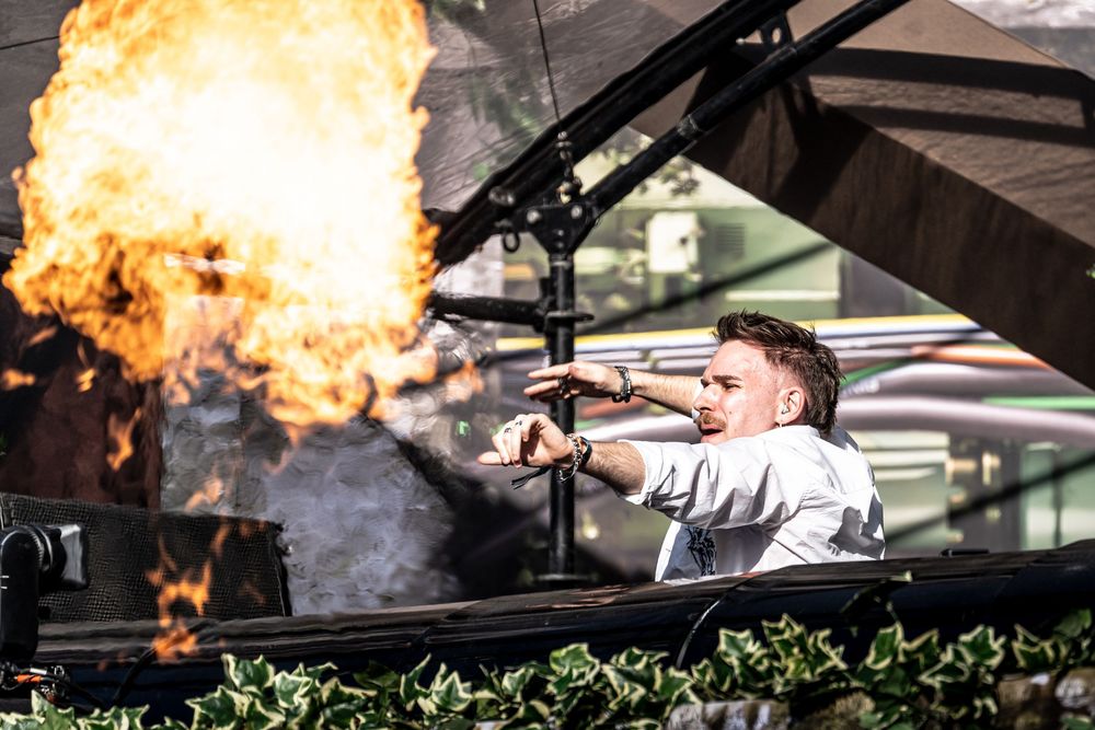 DJ performing on stage with dramatic fire explosion pyrotechnics behind him during live music festival event.