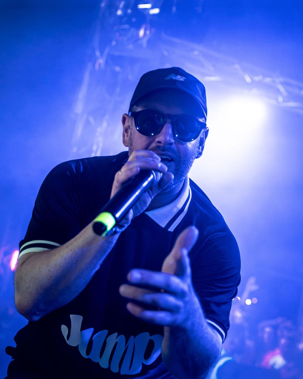 Male rapper performing on stage under blue lights, wearing sunglasses and dark cap, holding microphone close.